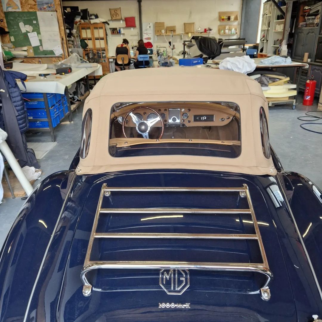 Arrière d'une MG MGA 1600 Mk II bleue avec capote beige et porte-bagages chromé dans un atelier de sellerie.