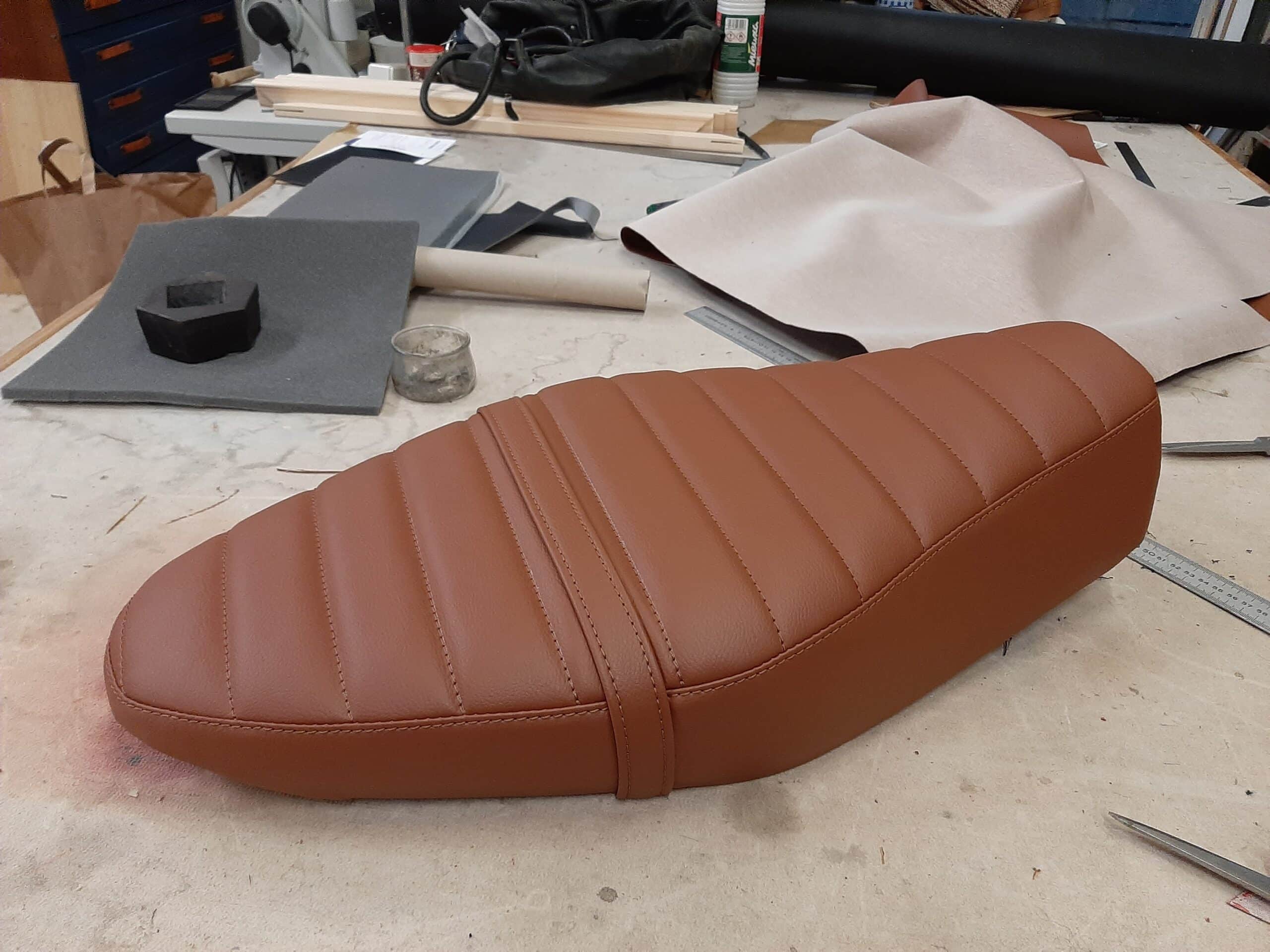 Selle de moto en cuir marron côtelée avec sangle centrale, posée sur un établi de sellerie artisanale.
