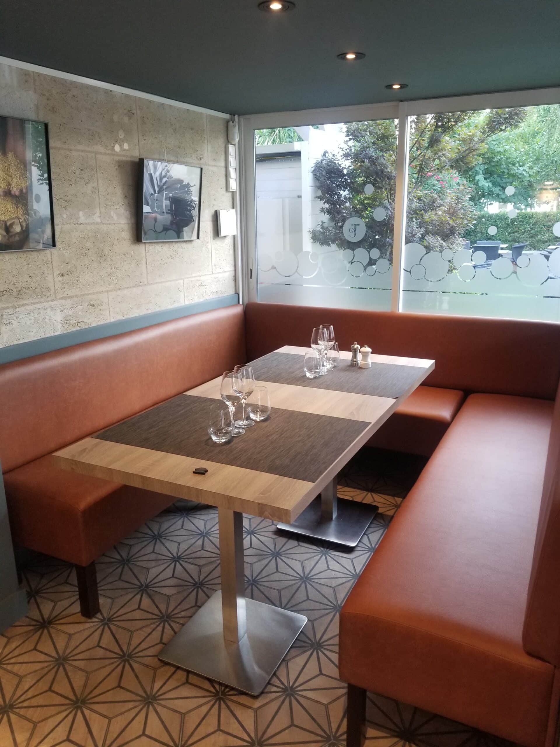 Banquette en cuir marron et table dressée dans un restaurant moderne avec sol géométrique et vue sur jardin.