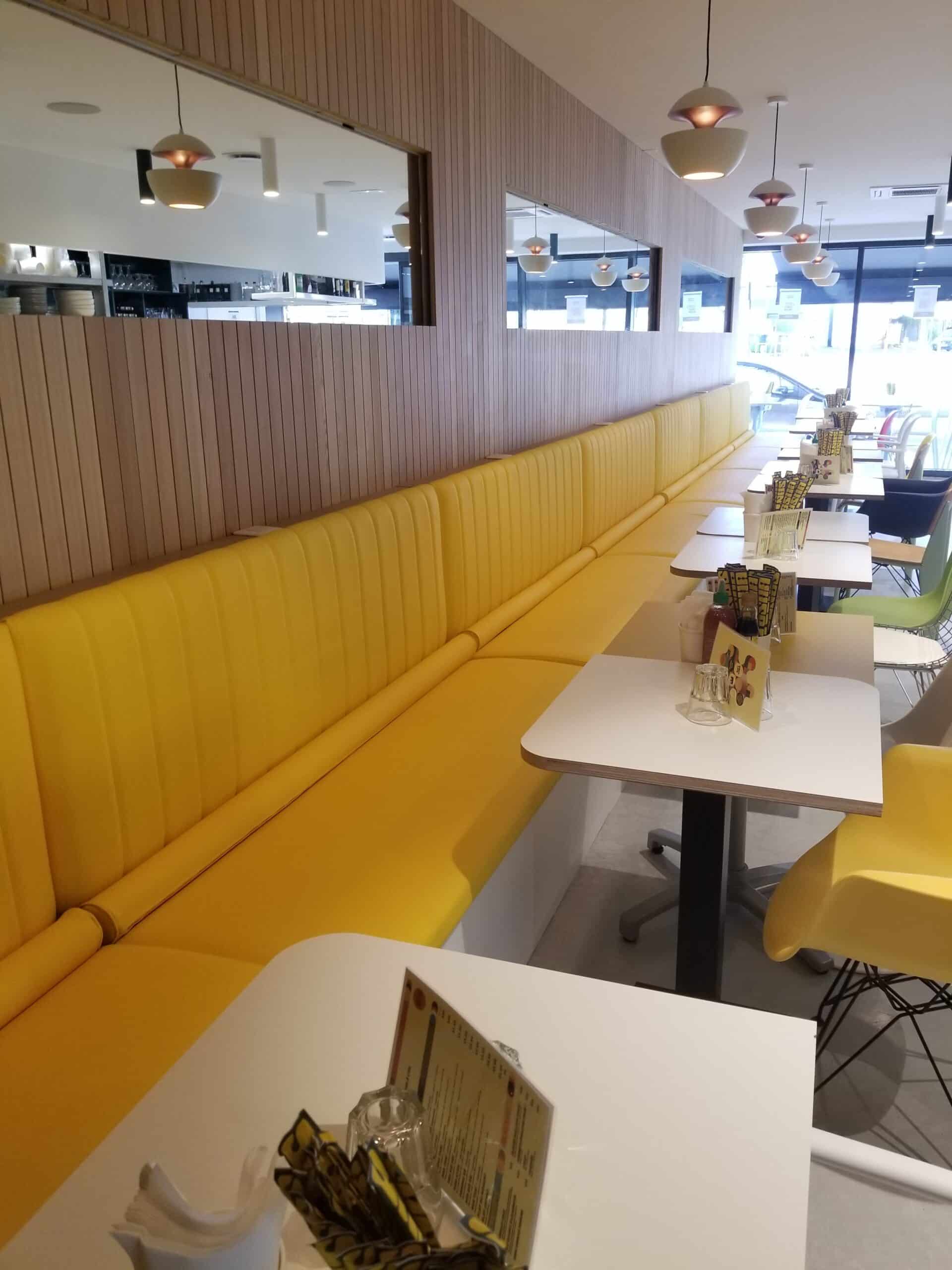 Intérieur de restaurant moderne avec banquette jaune, tables blanches et mur en lattes de bois.