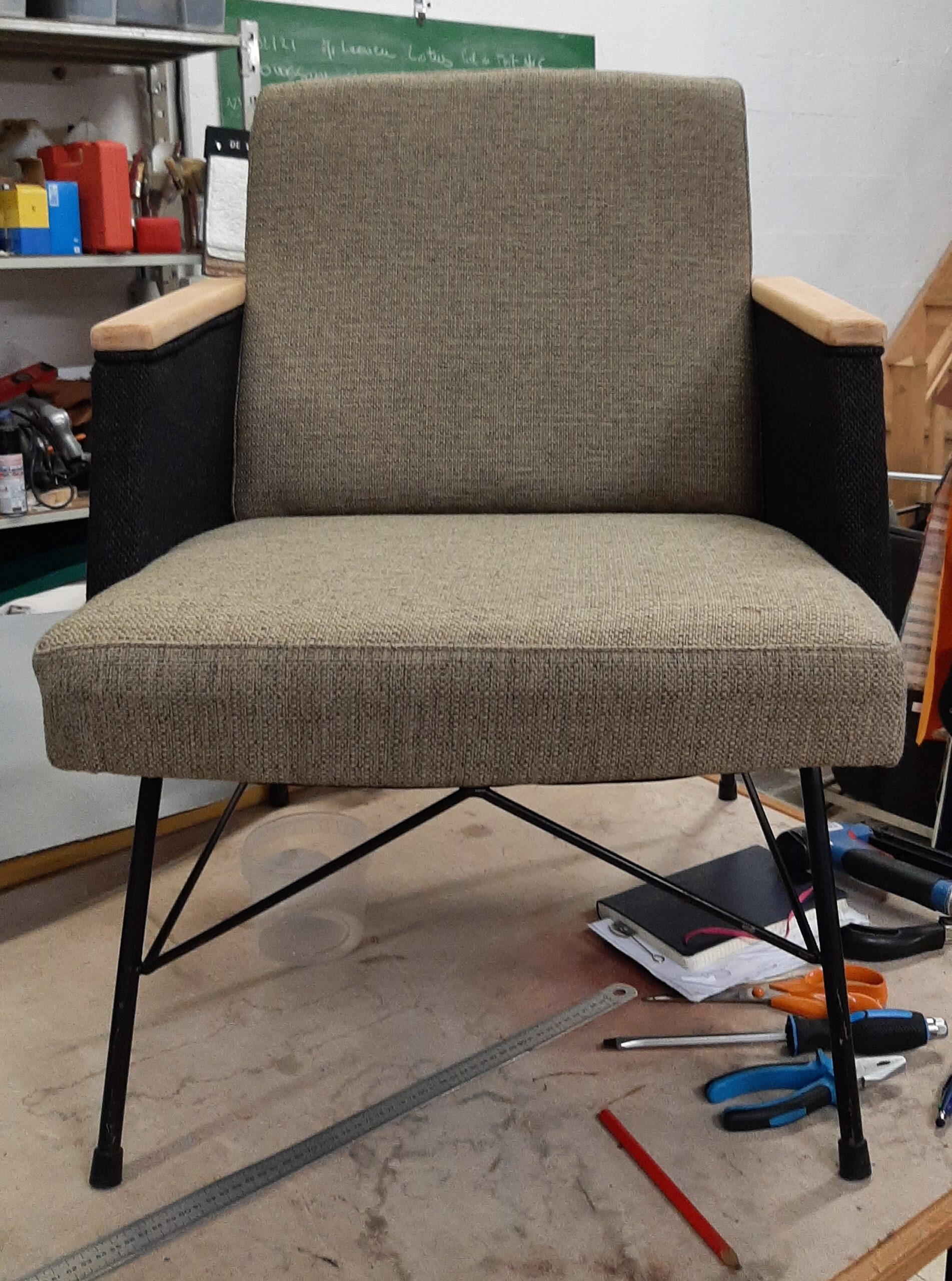 Fauteuil vintage restauré avec tissu beige texturé, accoudoirs bois et pieds en métal noir dans un atelier.